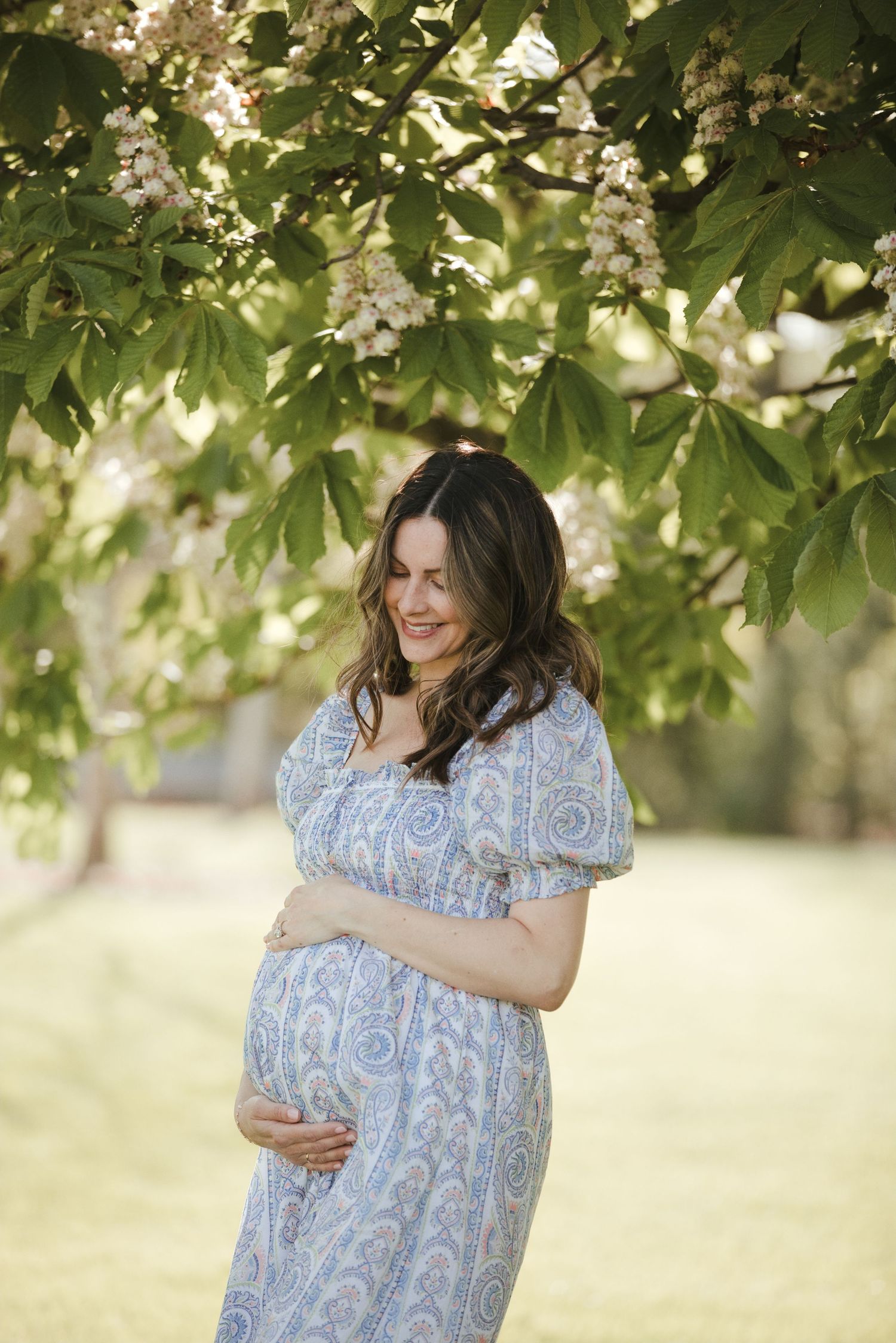 Pregnant woman in blue floral dress cradling baby bump under blooming tree in sunny park.