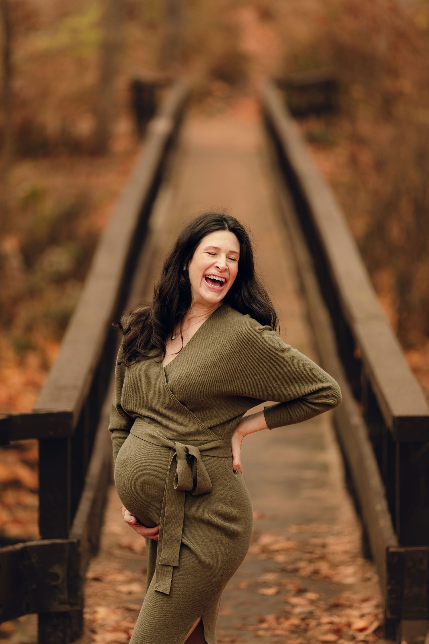 Maternity photos on wooden bridge with olive dress against autumn backdrop.