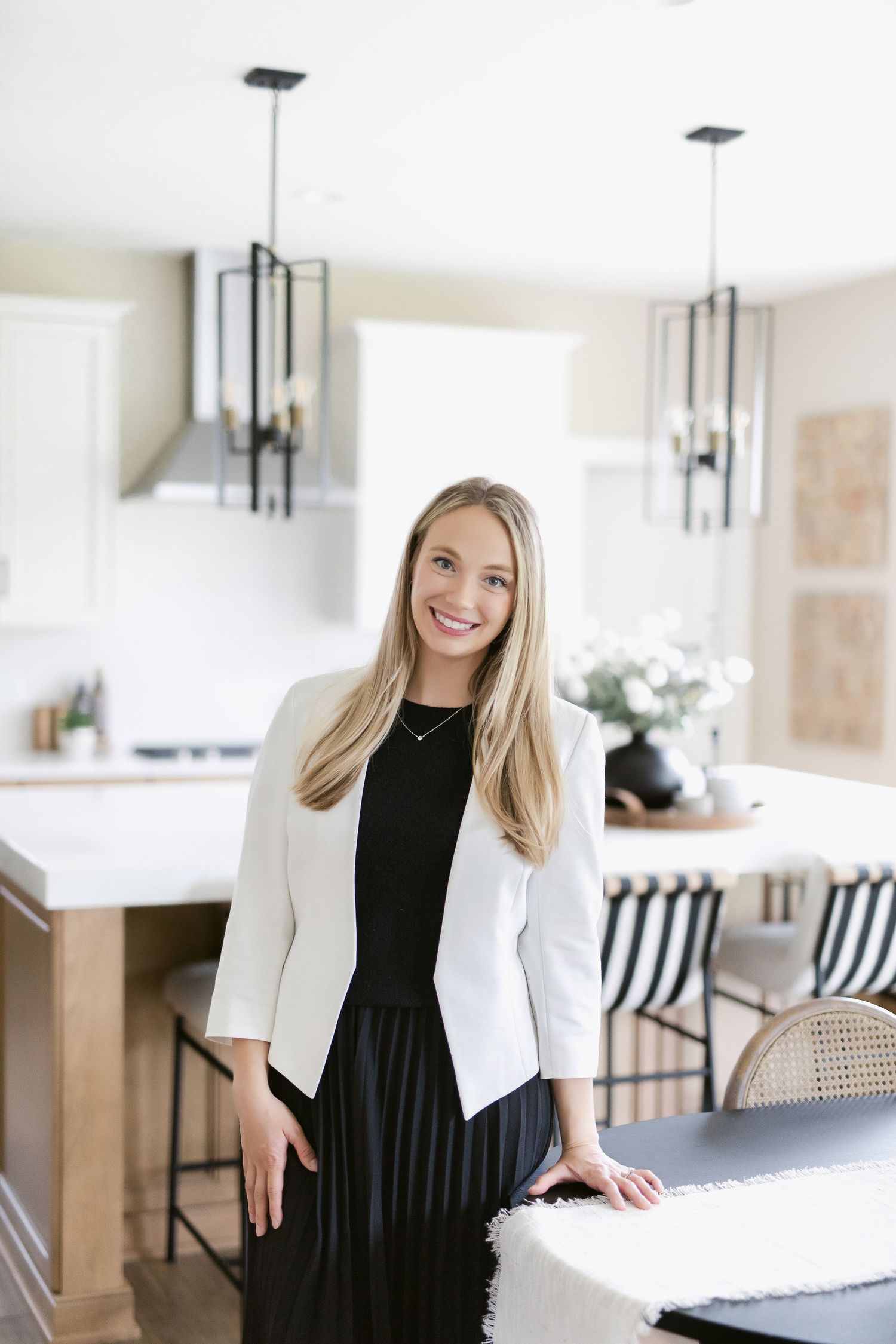 Professional in white blazer and black dress poses in modern kitchen with pendant lights.