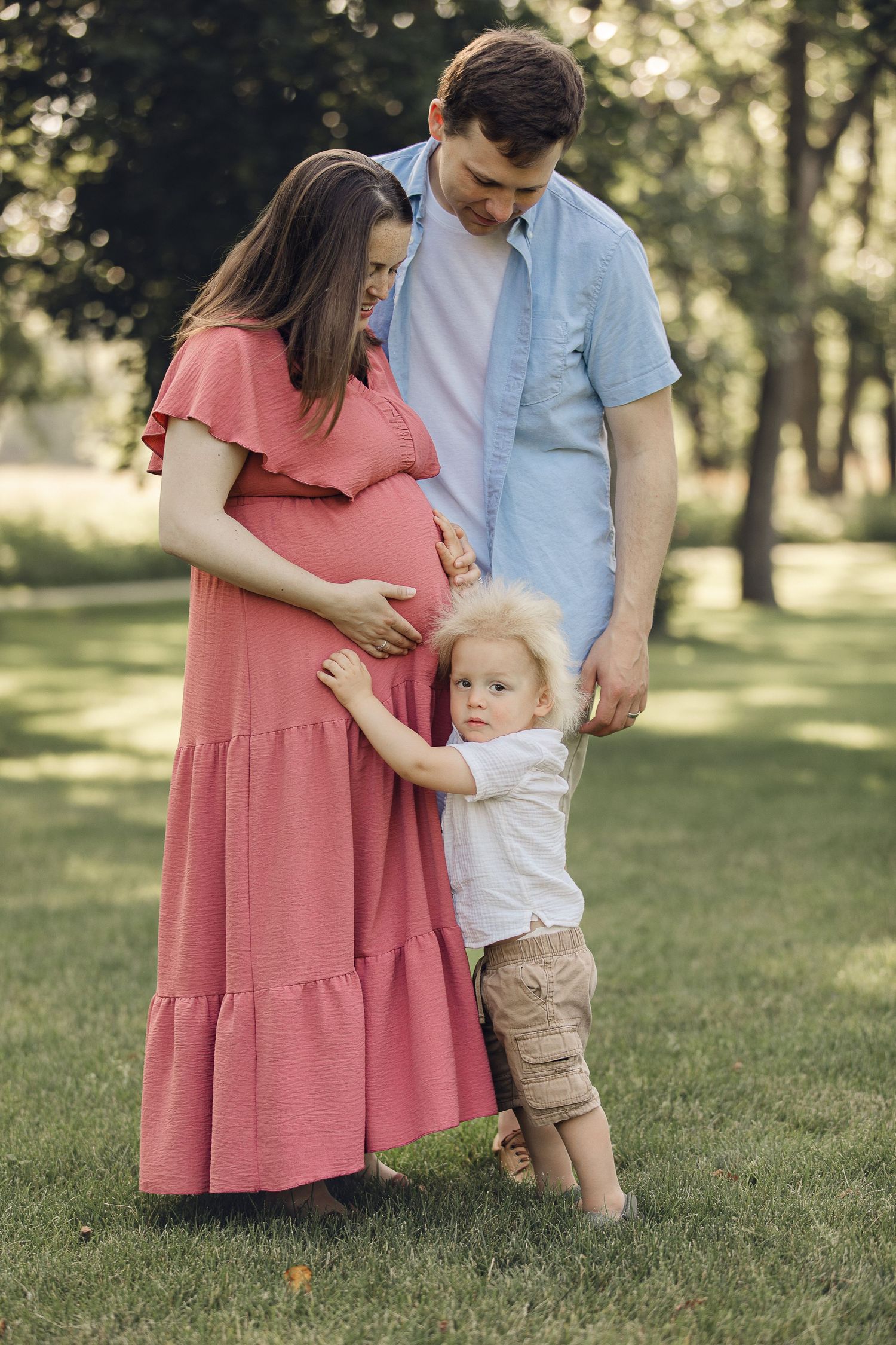 A family poses outdoors in a park with a pregnant woman in coral dress and toddler holding her belly.
