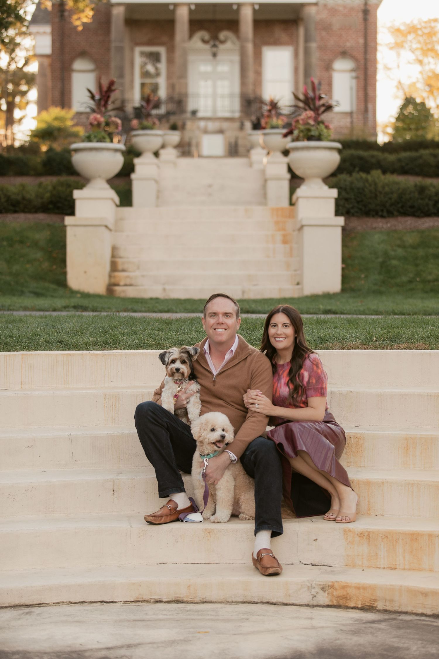 A couple sits with their two dogs on stone steps in front of a stately home.