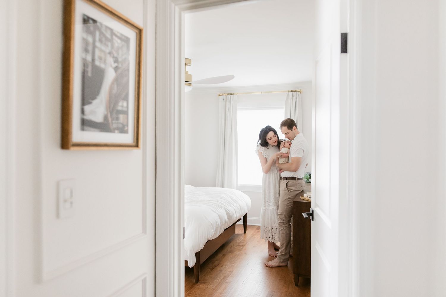 View through doorway into bright bedroom with hardwood floors and white walls showing two people standing in soft light.