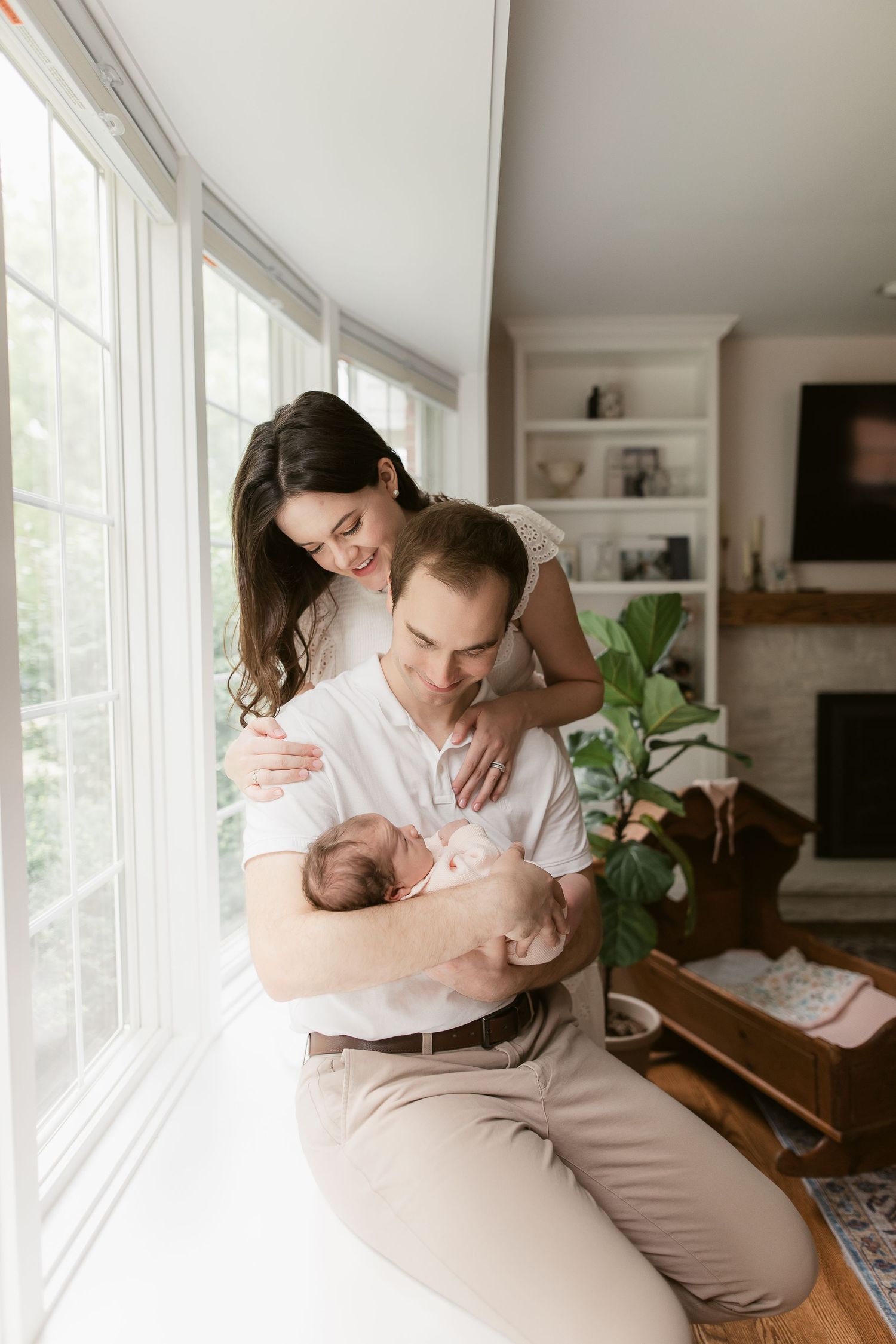 Family cuddles on couch near large windows with indoor plants in background.