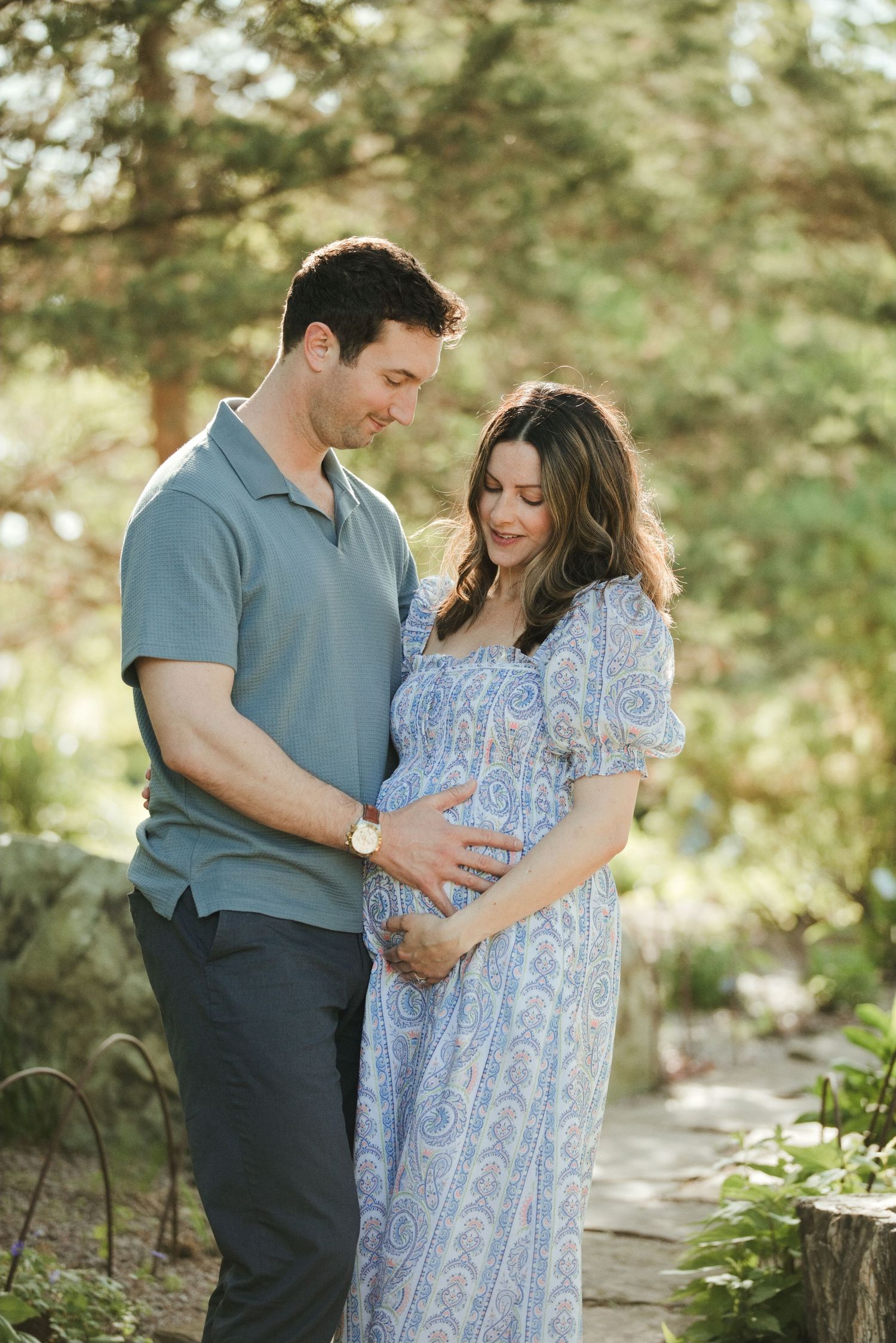 Couple tenderly cradling pregnant belly outdoors in garden, wearing blue outfits, golden hour light filtering through trees.