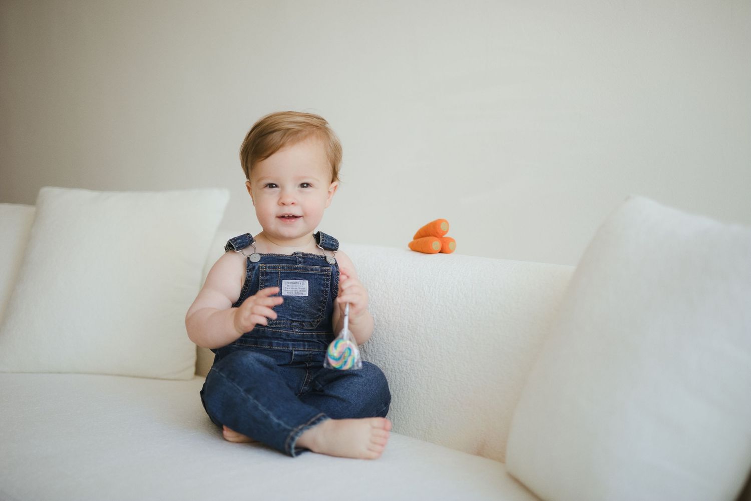 Adorable baby boy in denim overalls sitting on a white couch, smiling cheerfully.