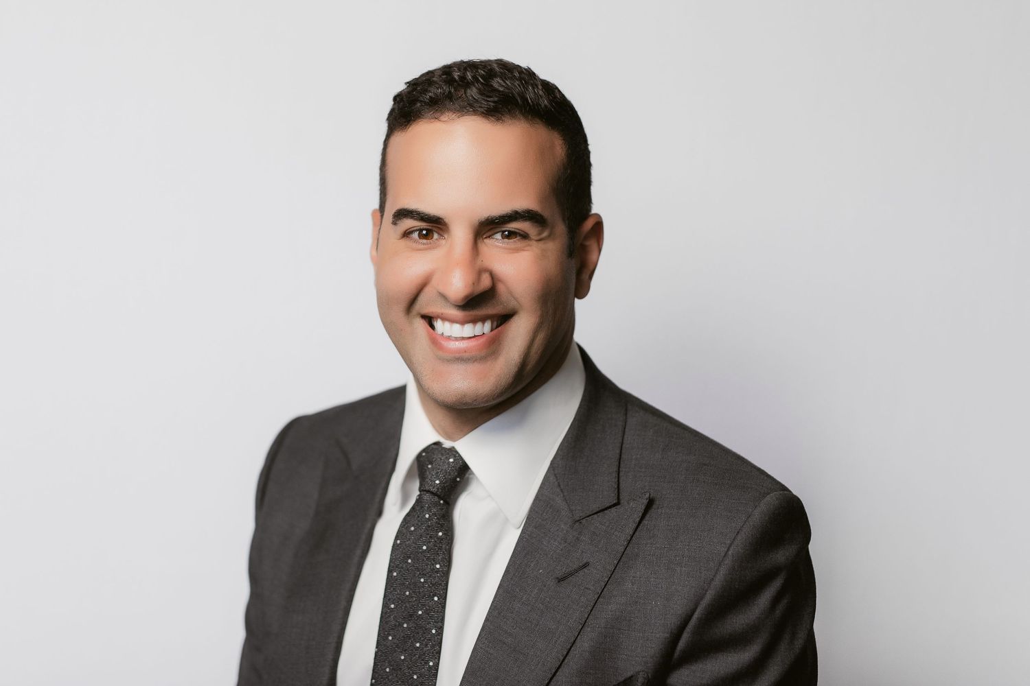 Professional headshot of someone in a gray suit and polka dot tie smiling against white background.