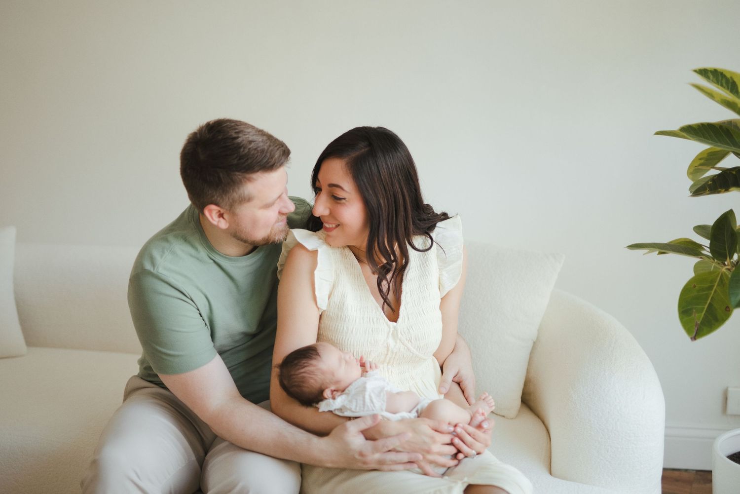 A couple sits on a white sofa, smiling at each other while cradling their newborn baby in a bright, cozy room.