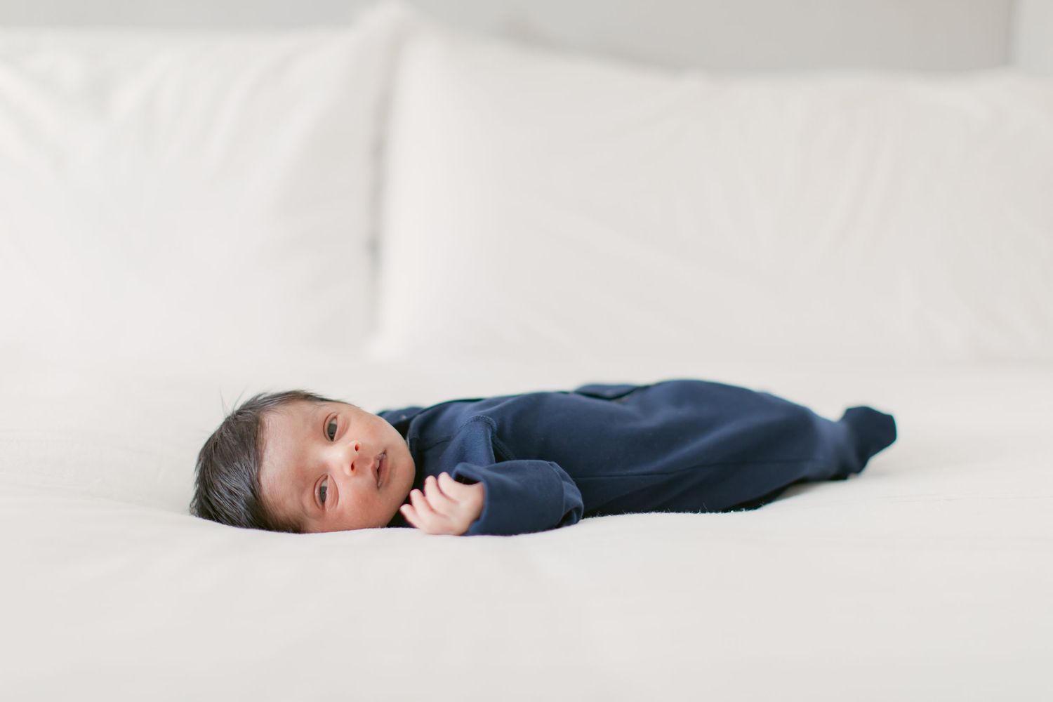 Newborn baby in navy blue sleeper lying on white bedding, looking at camera during professional infant photography session.