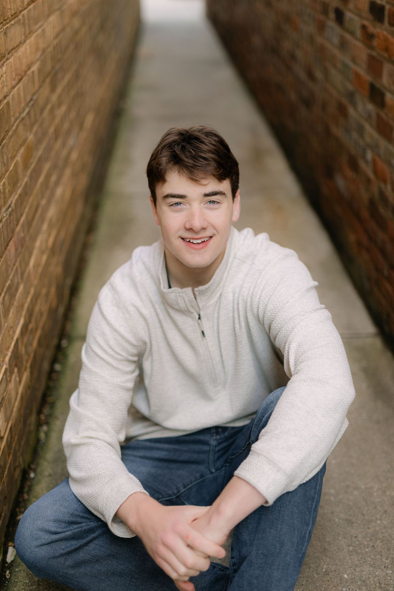 Young man in white quarter-zip sweater and jeans sitting and smiling in a narrow brick alleyway.