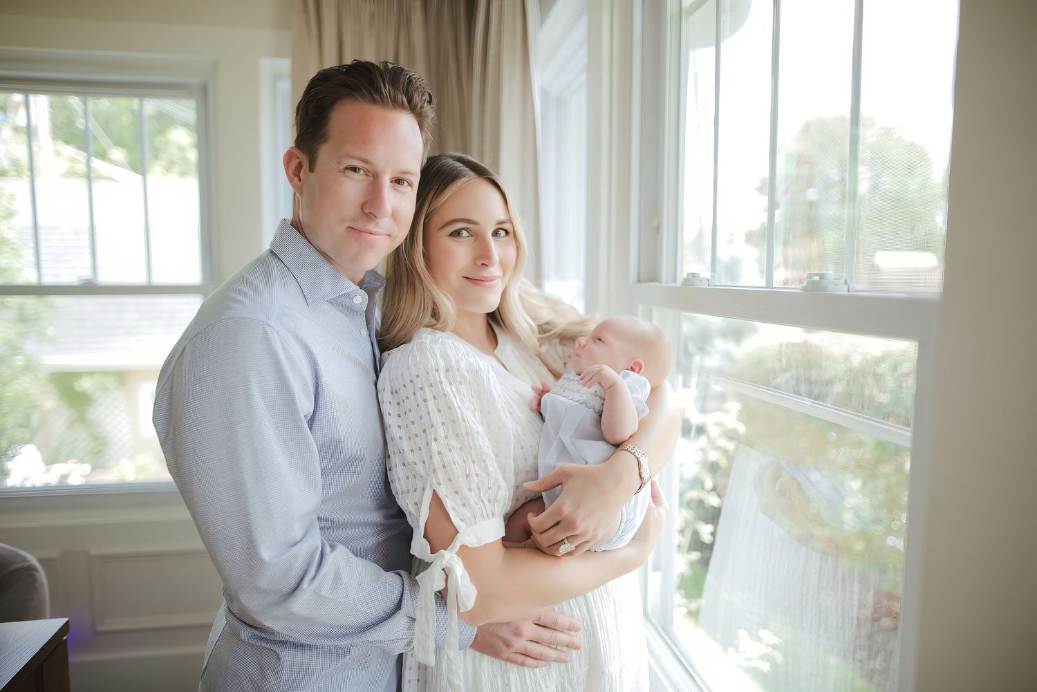 A smiling couple poses indoors near a window, the woman holding a newborn baby wrapped in a soft blanket.