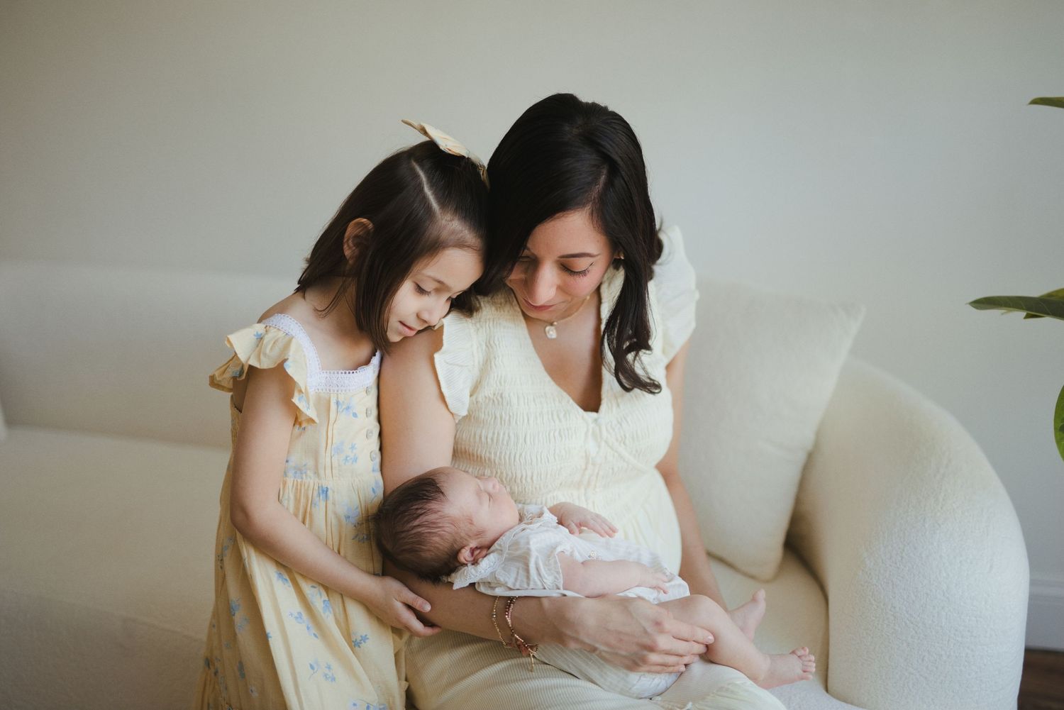 A mother in yellow holds her newborn while her young daughter in a floral dress looks on lovingly indoors.
