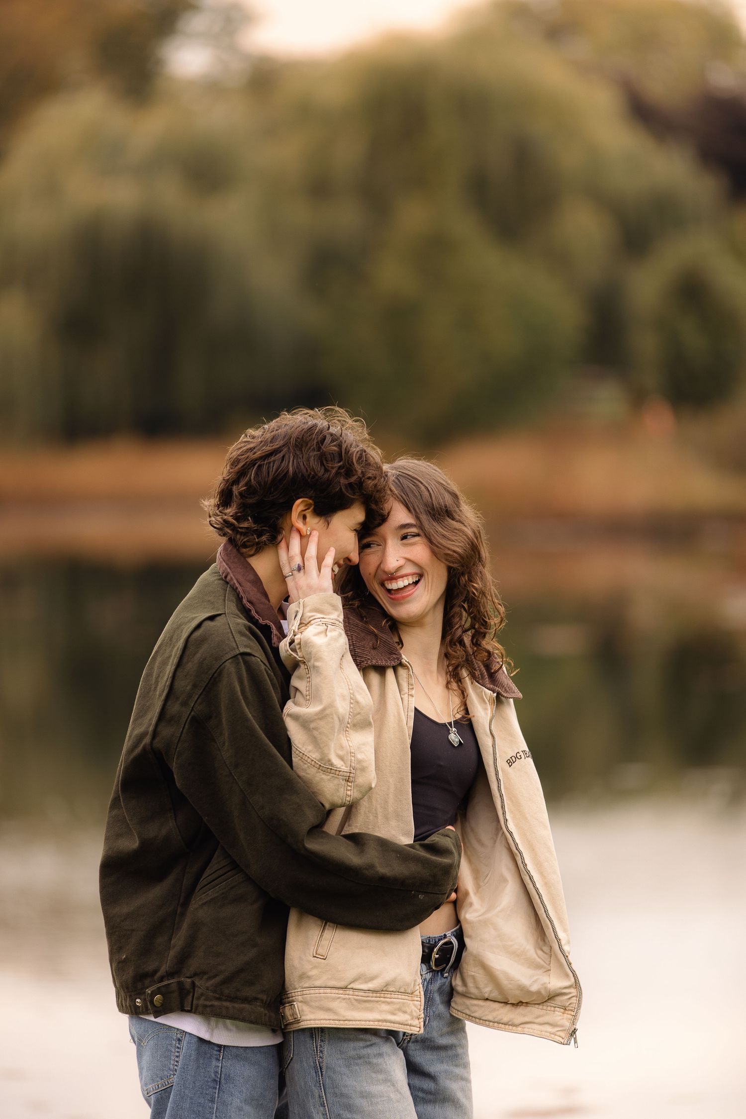 Couple laughing together by a lake during golden hour with autumn foliage in the background.