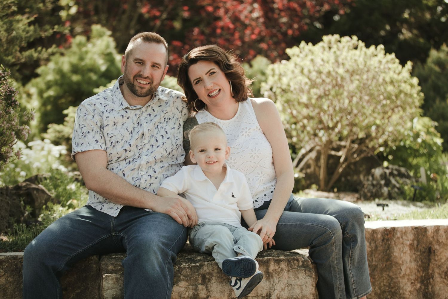 A happy family of three sits outdoors on a stone wall, with lush green gardens in the background.