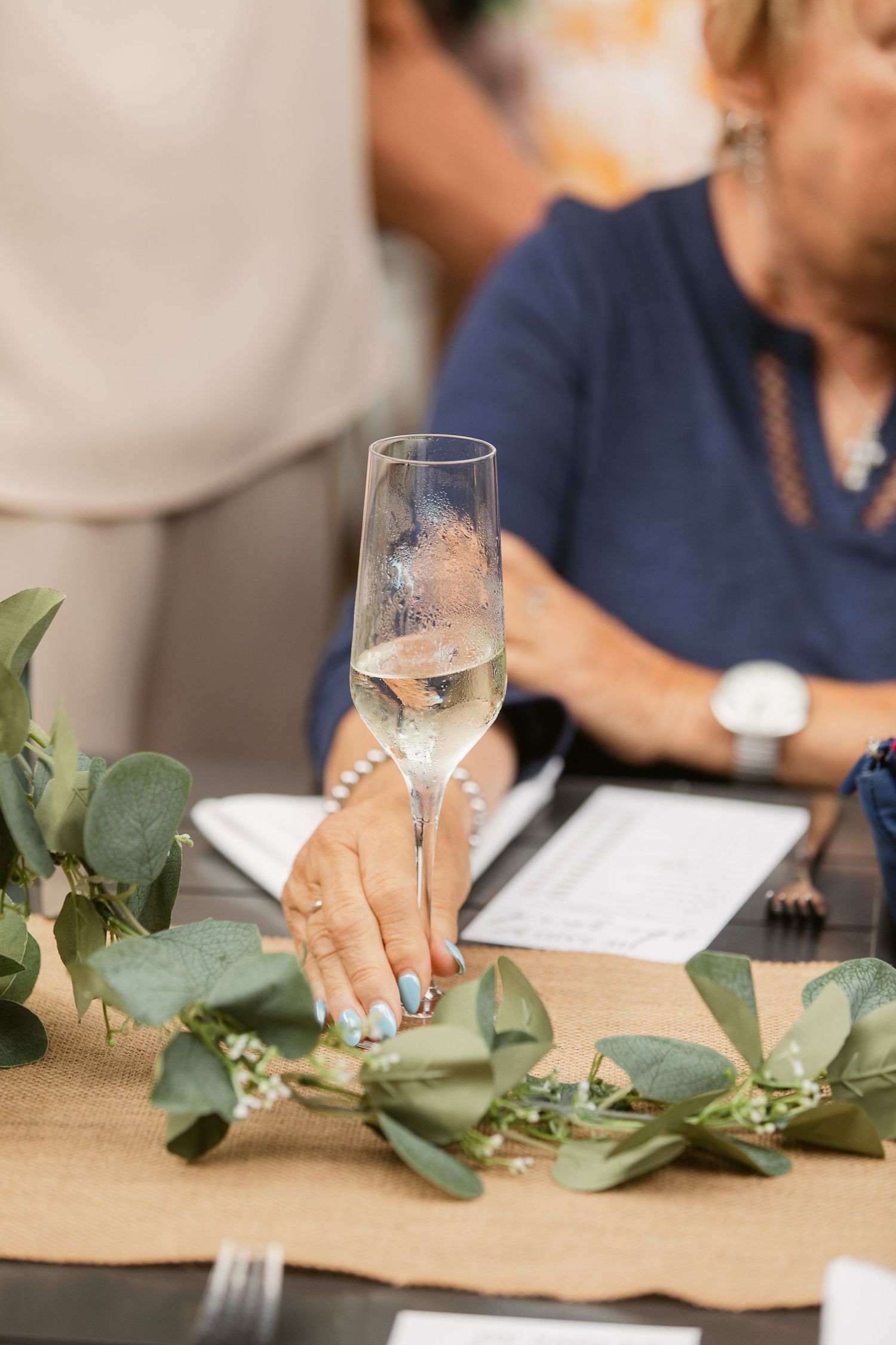 Hand holding champagne flute at elegant wedding reception table with eucalyptus garland centerpiece.