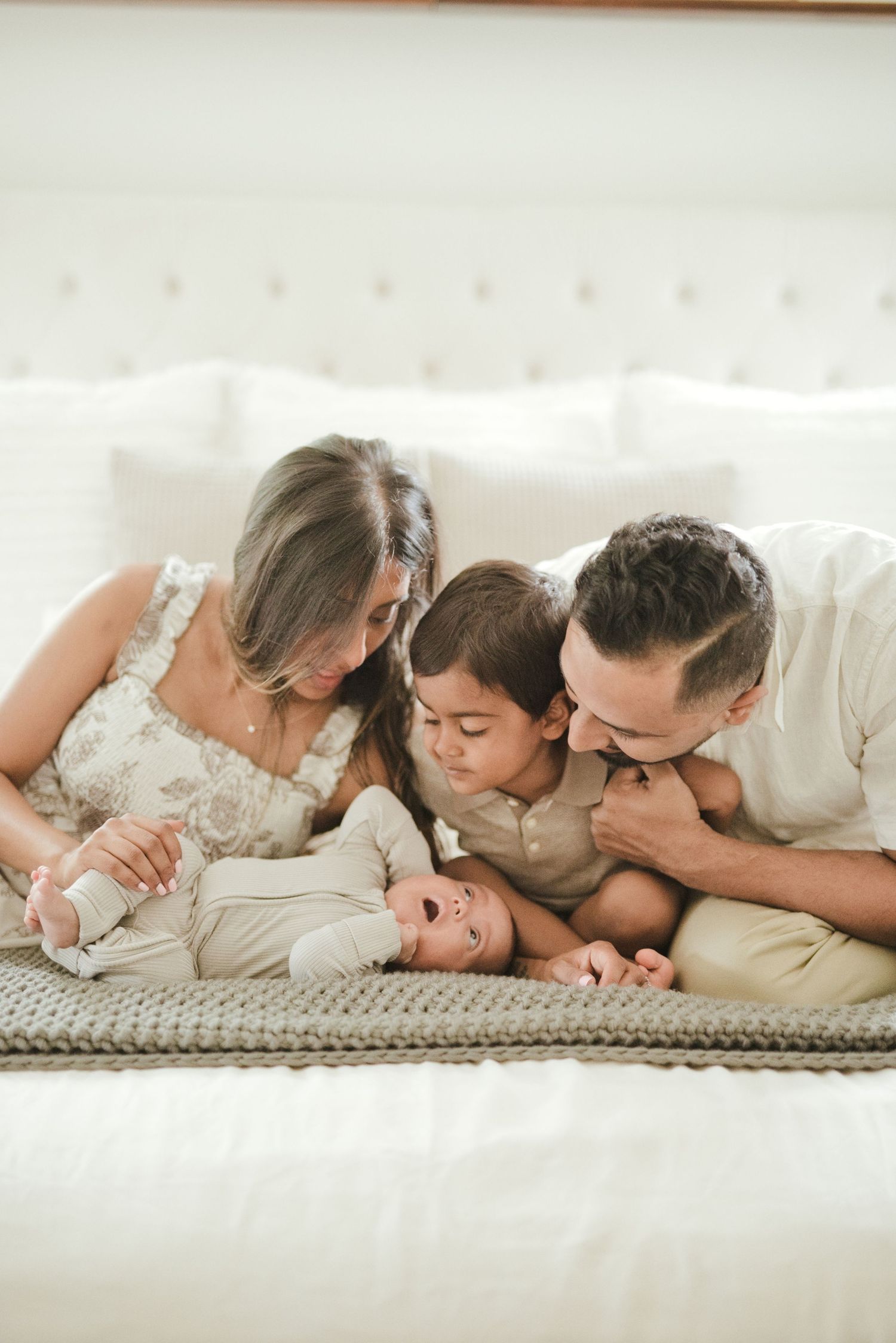 Family of four on bed, parents and toddler smiling at laughing newborn baby in cozy white bedroom.