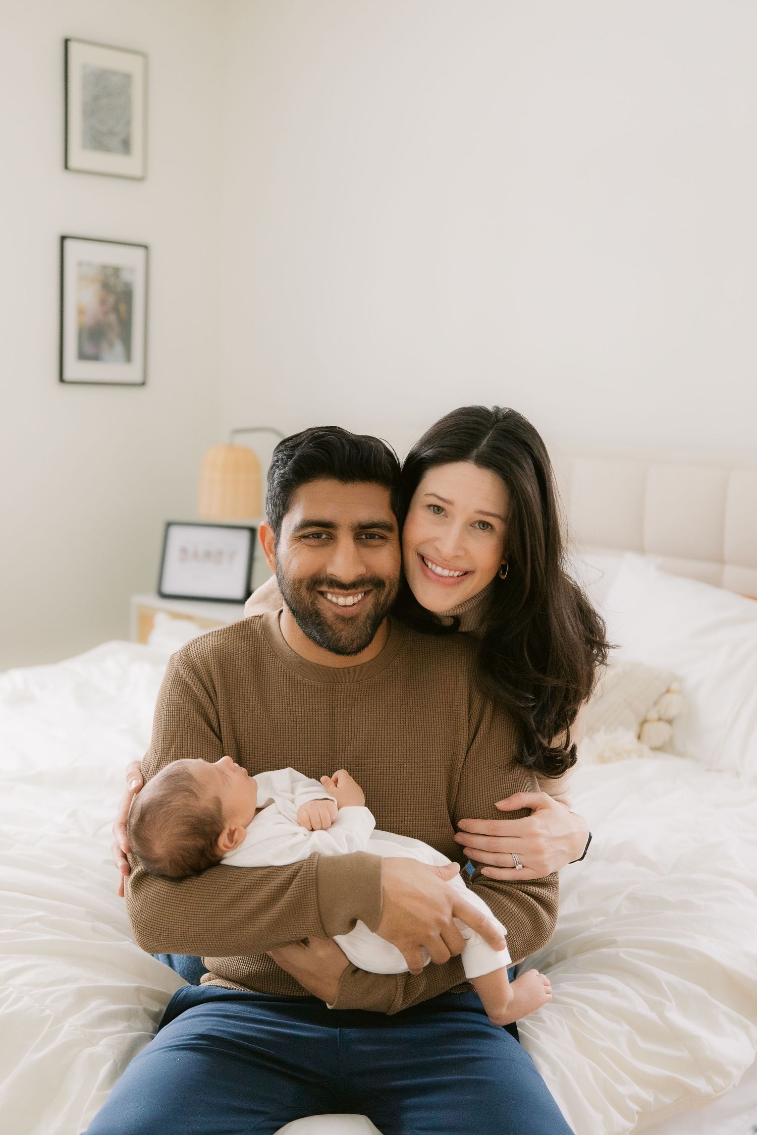 A couple sits together on a white bed while holding and admiring their newborn baby.