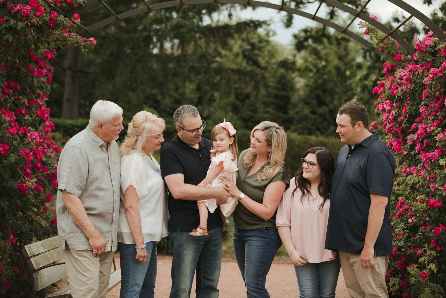 Multi-generational family gathering under floral arch with pink roses, sharing joyful moment with baby outdoors.