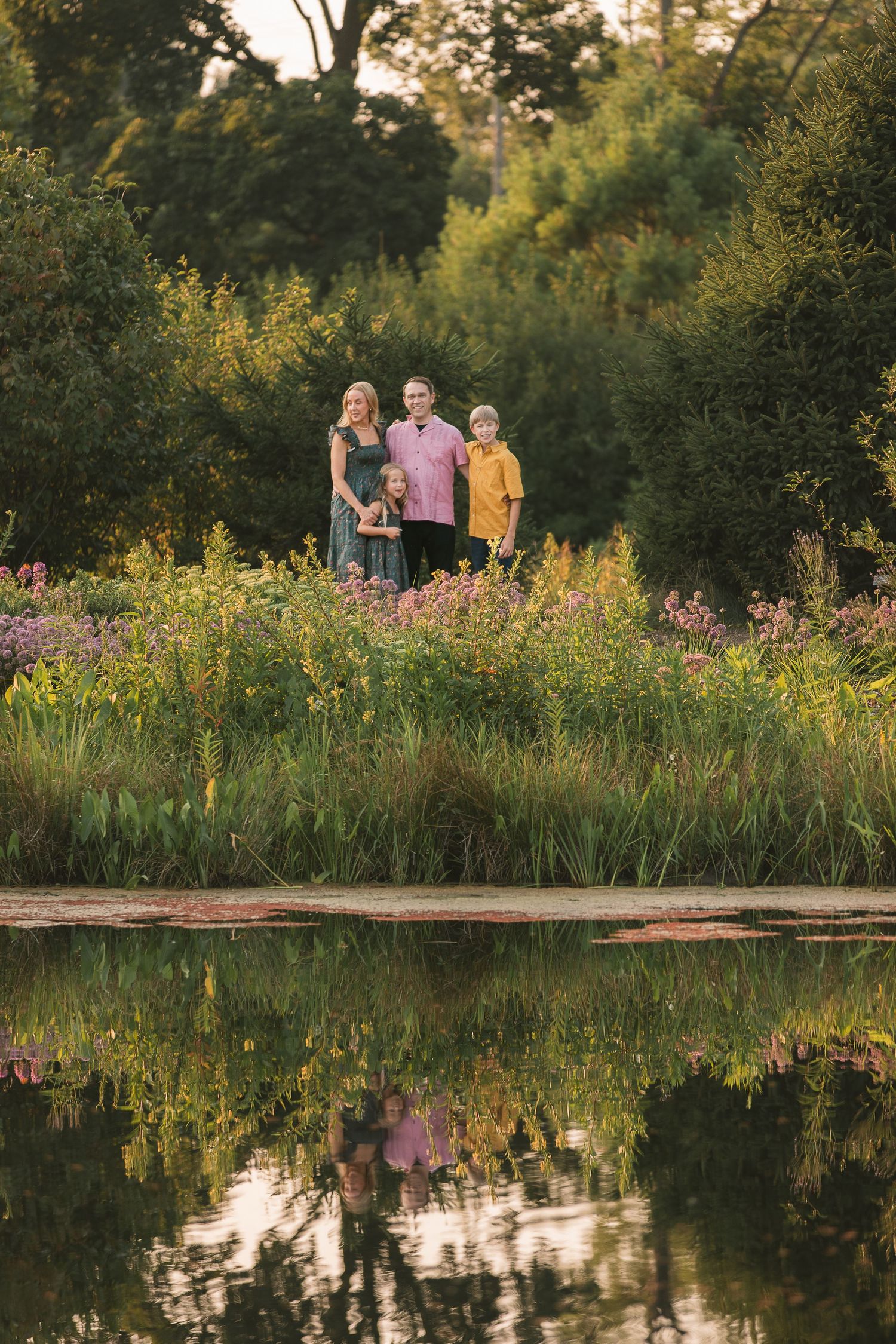 Family of four standing among wildflowers beside a reflective pond, surrounded by lush green trees at golden hour.
