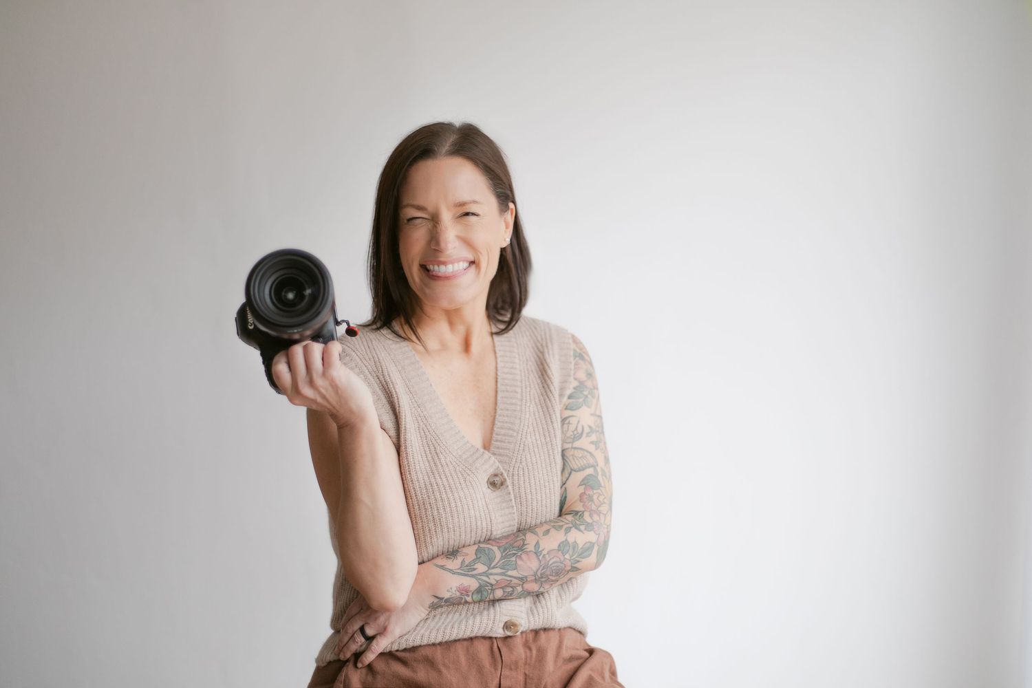 Smiling woman with tattoos holding a camera, wearing a beige cardigan and rust pants against a white background.