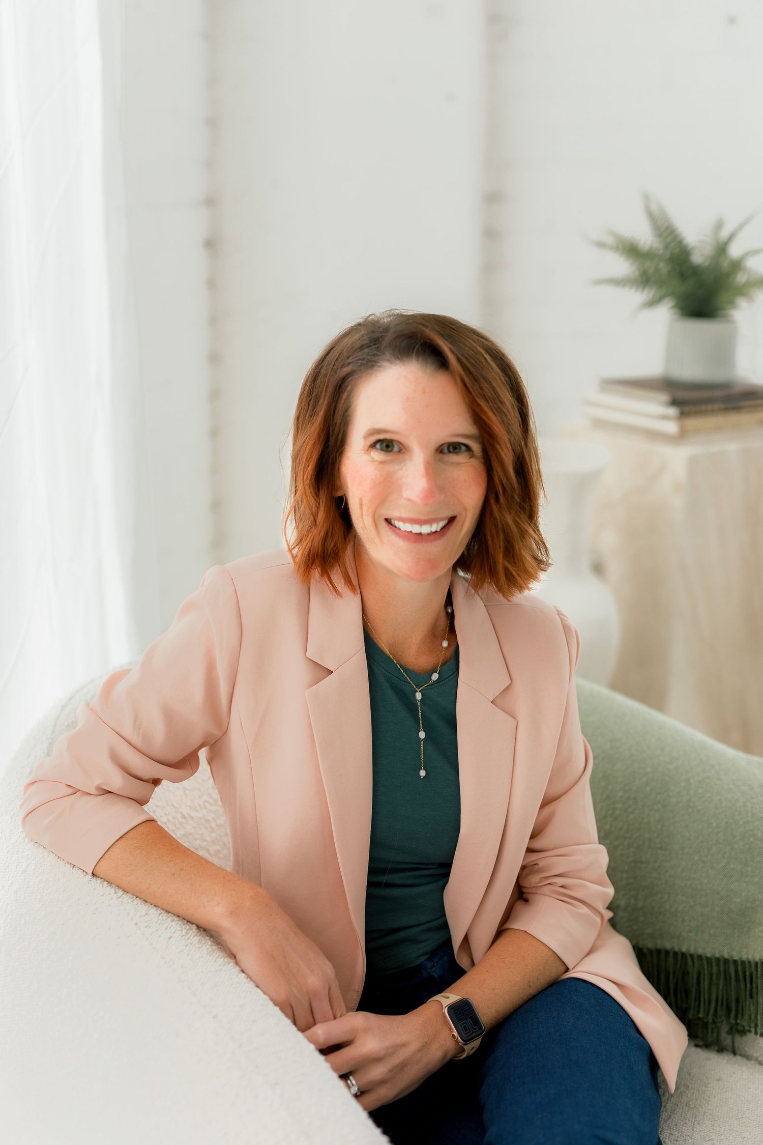Professional portrait wearing pink blazer and dark top seated on white couch.