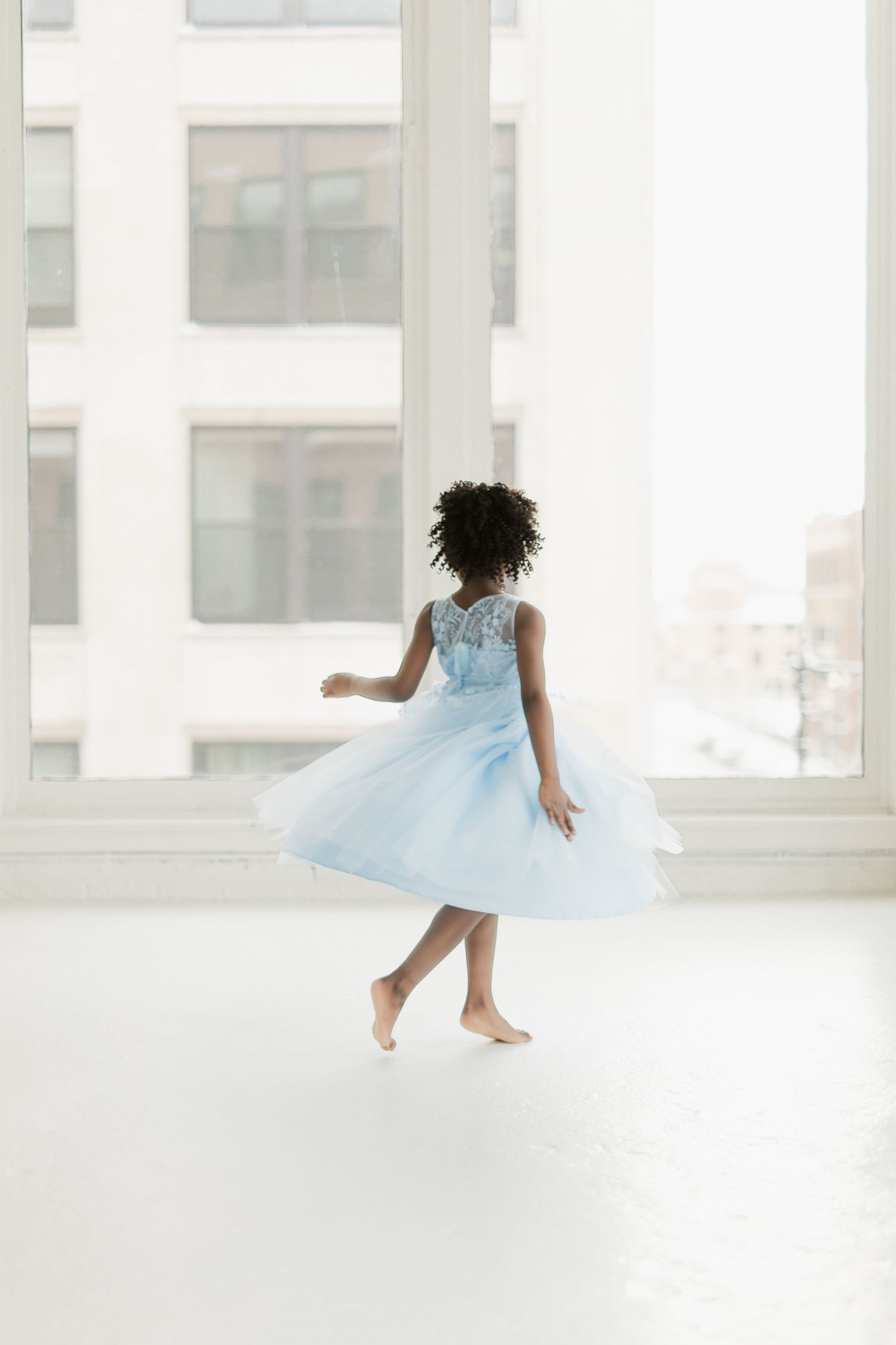 Young girl in a light blue tulle dress twirling barefoot in a bright white studio near large windows.