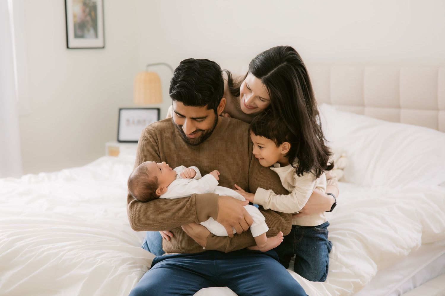 A family sits together on a white bed sharing tender moments with their newborn baby in a bright room.
