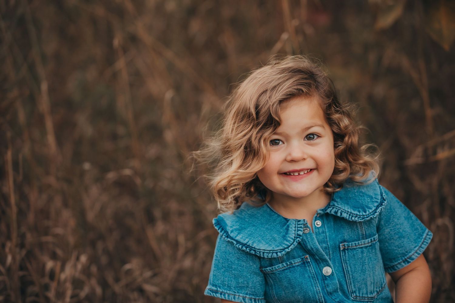 Child with curly hair wearing a blue denim top smiles brightly against a blurred natural background.
