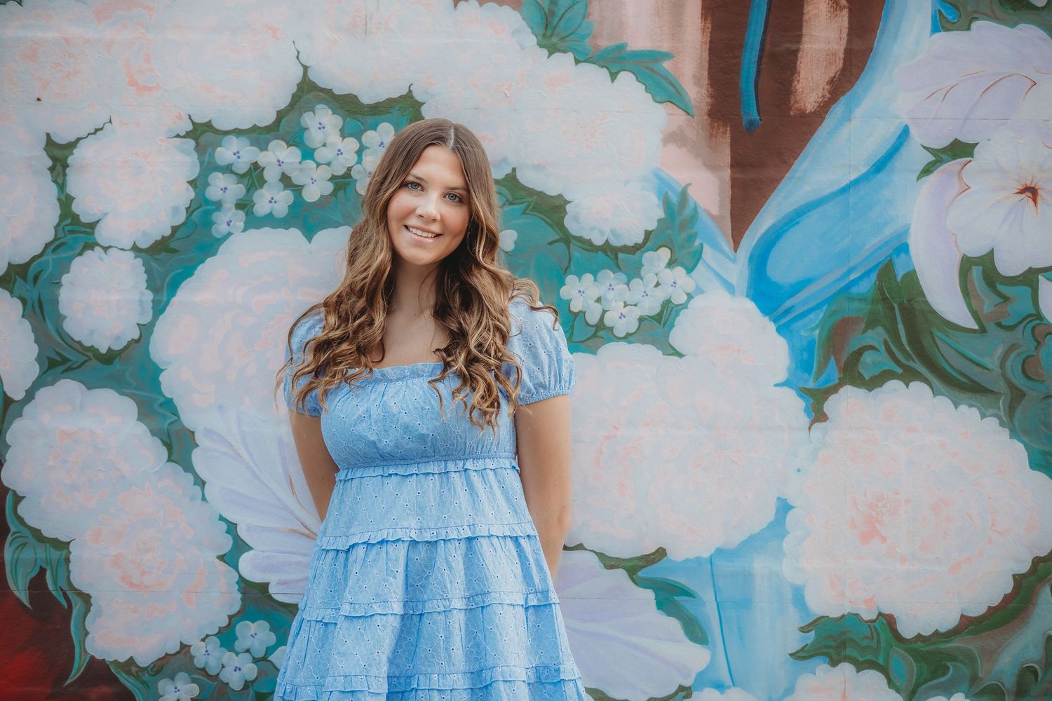 Young woman in a blue tiered dress smiling in front of a floral mural with white flowers.