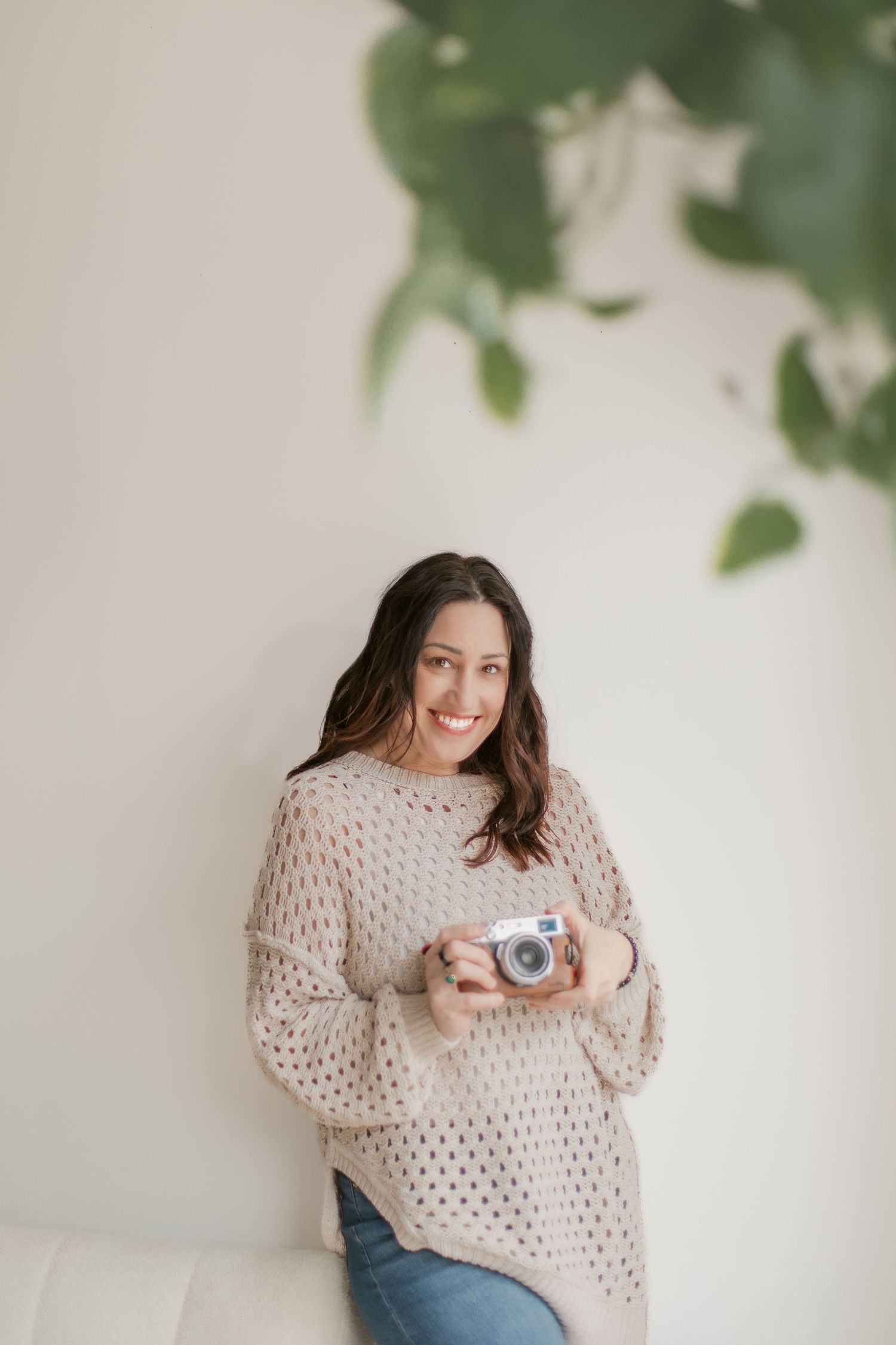 Person in cozy beige sweater and jeans poses with vintage camera against white wall with green foliage.