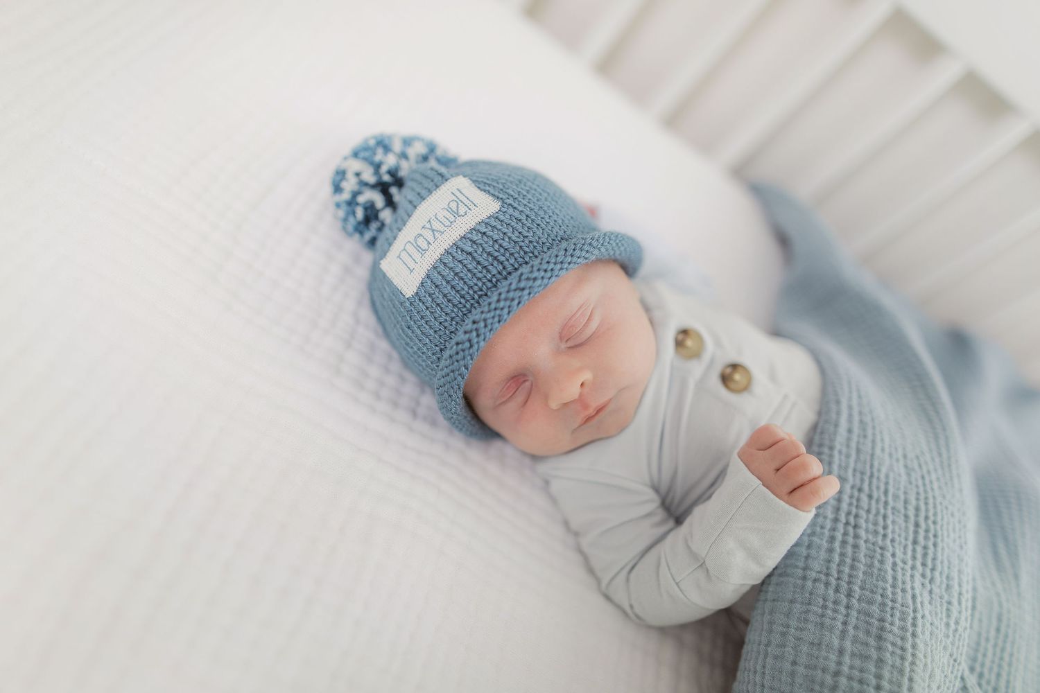 Sleeping newborn wearing soft blue knitted hat and gray onesie lying on white bedding.