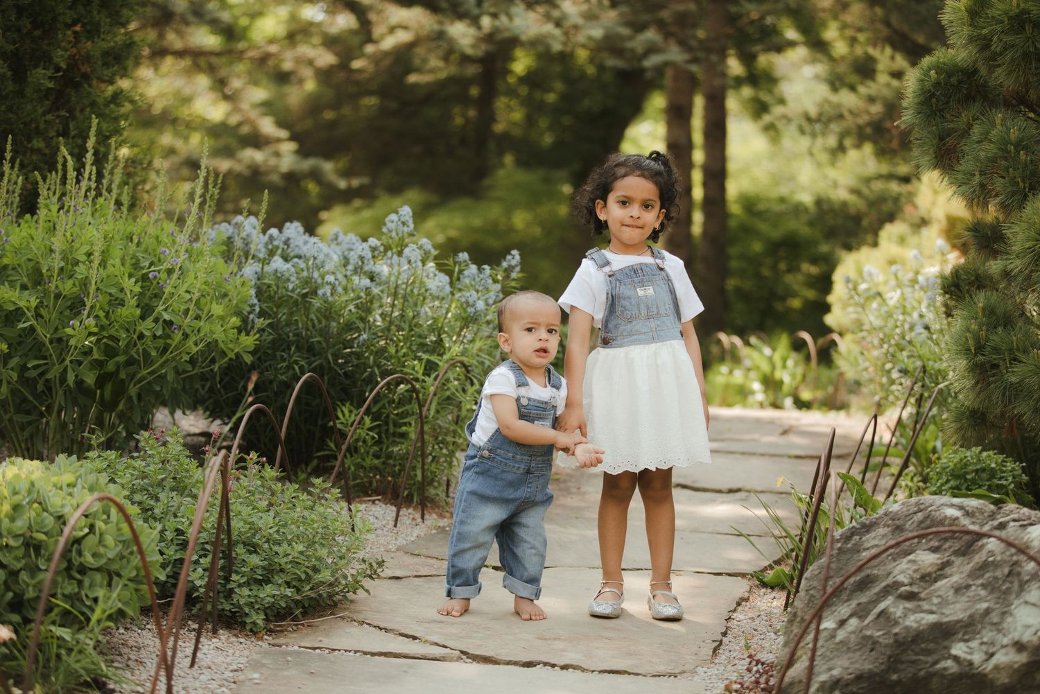 Two young siblings in a lush garden, toddler boy in overalls and girl in white dress on a stone path.