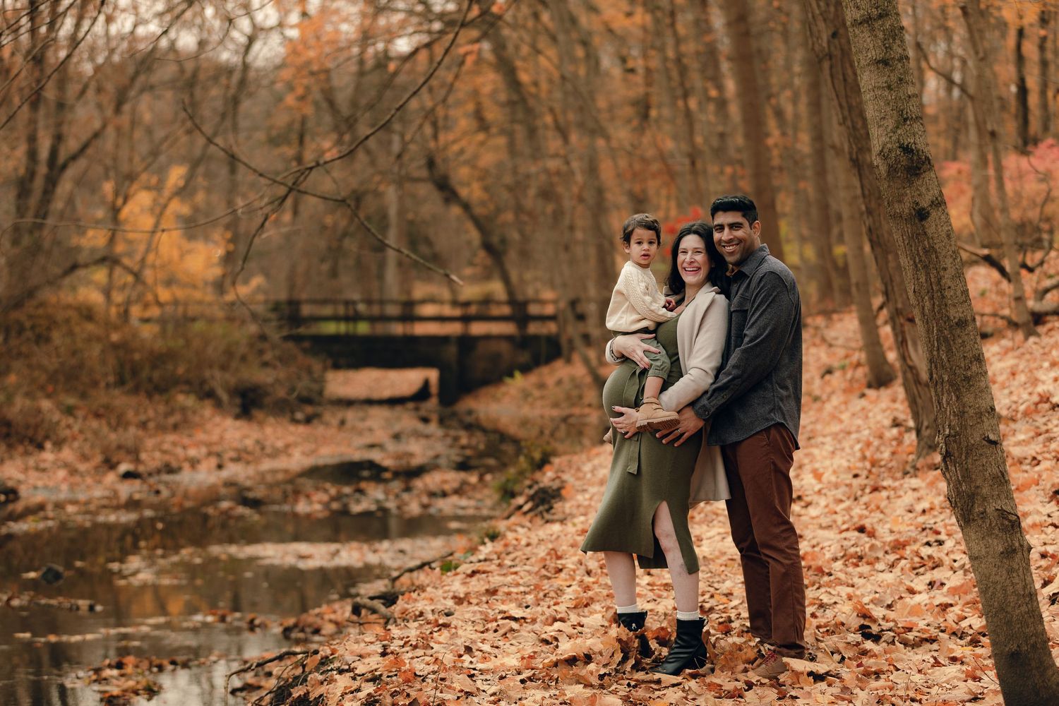 Family of three poses among autumn leaves beside a creek in a wooded park, with a bridge visible in background.