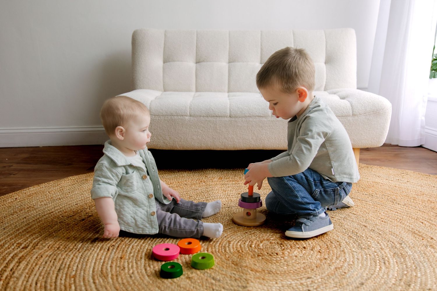 Two young children sit on a round jute rug playing with toys in front of a white tufted couch.