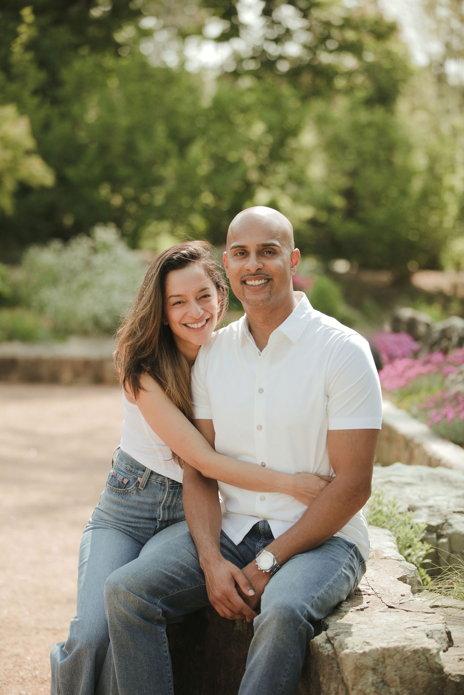 A smiling couple sits together outdoors in a garden, wearing white tops and jeans, surrounded by lush greenery.