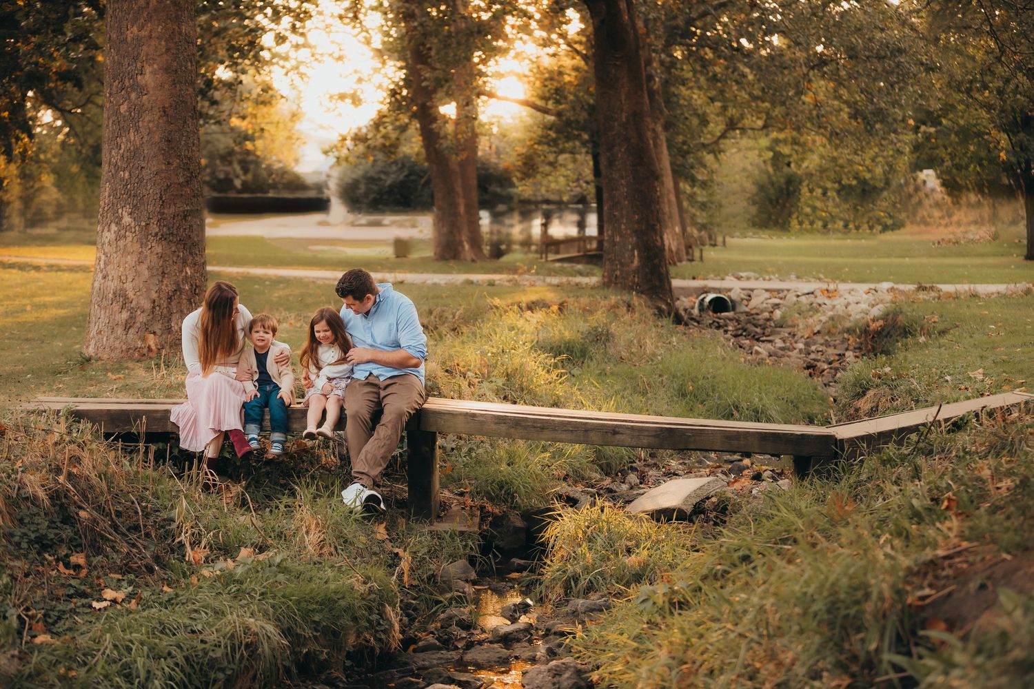 A family sits on a wooden footbridge in a peaceful park setting during golden hour.