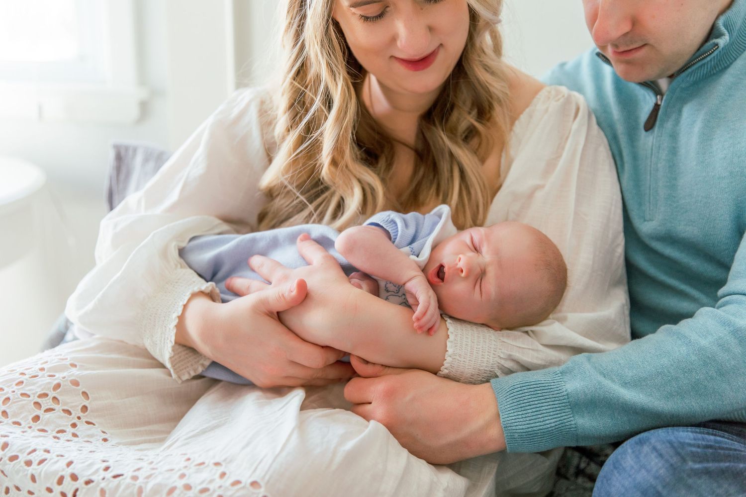Parents tenderly hold their sleeping newborn baby on a white bed.