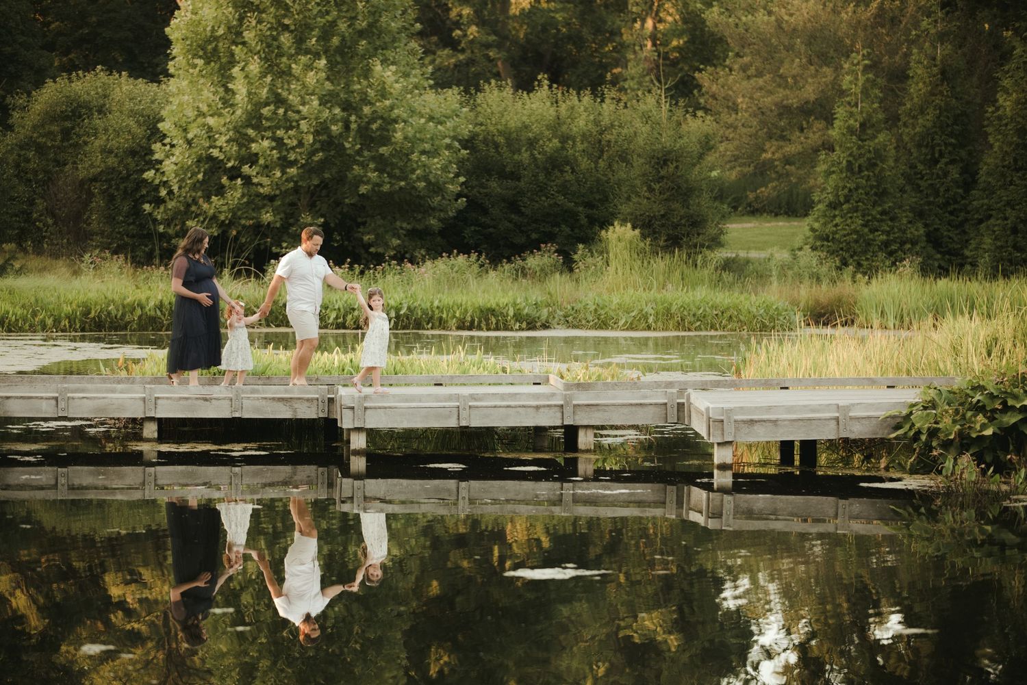 Family of three holding hands on a wooden dock surrounded by lush greenery reflected in calm water.