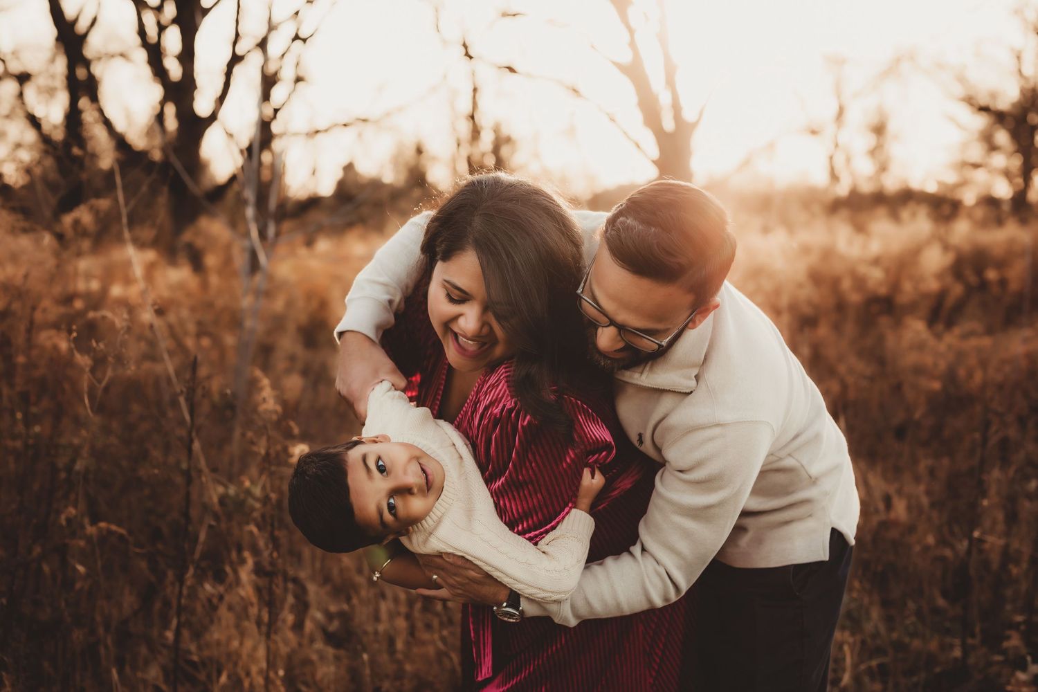 A couple embraces in a warm autumn field during golden hour as sunlight filters through bare trees.