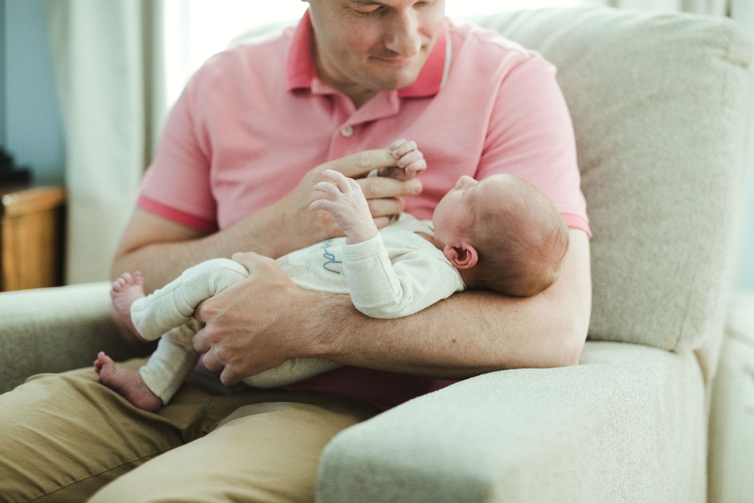 Father in pink shirt cradling newborn baby while sitting on beige sofa at home.