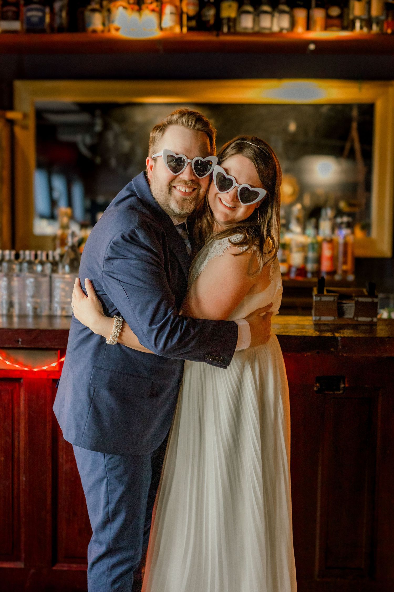 A couple in wedding attire hugs and smiles wearing white heart-shaped sunglasses in front of a bar.
