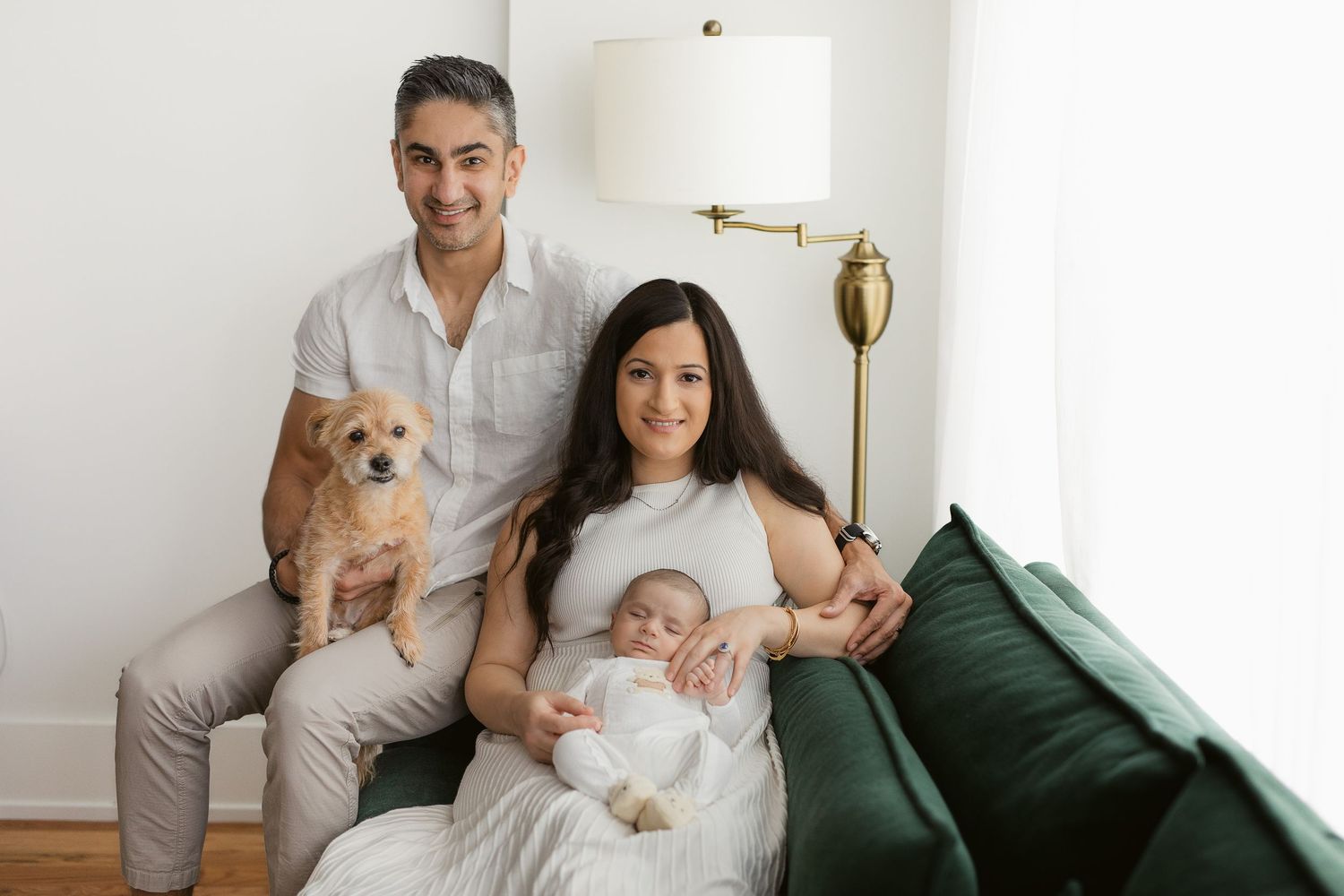 Family portrait on green couch with small dog and infant in bright living room.