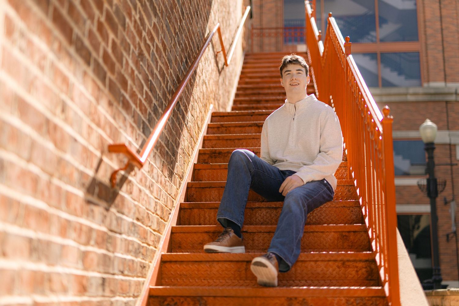 Young man sitting on vibrant orange outdoor stairs beside a brick wall in warm sunlight.
