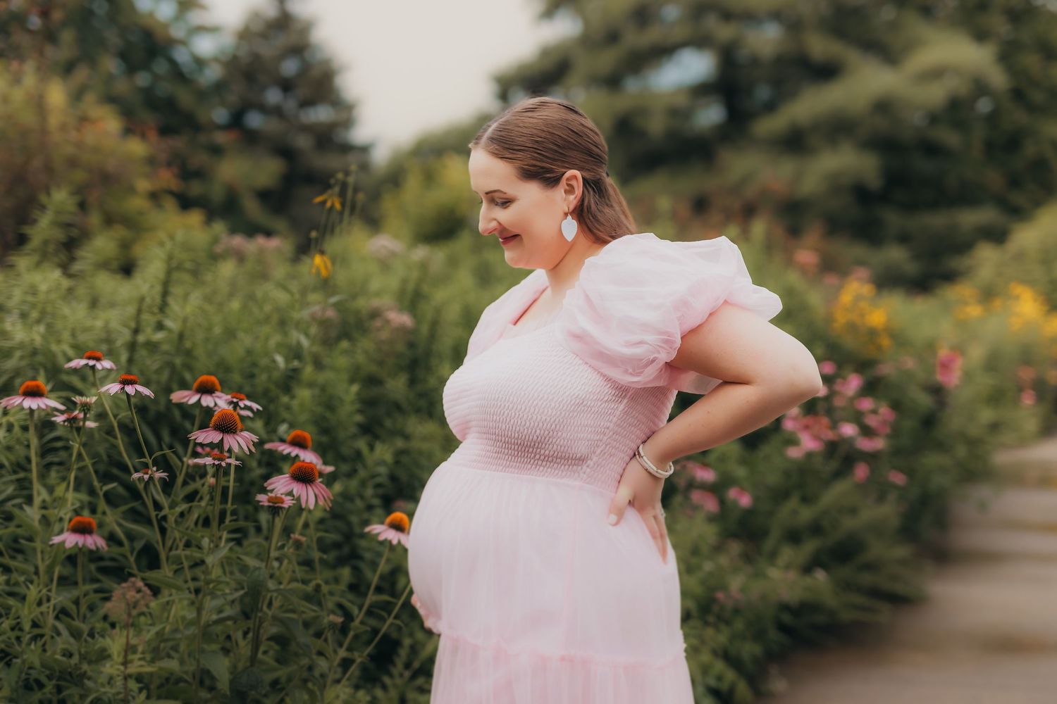 Pregnant woman in pink dress standing among colorful garden flowers, smiling down at her baby bump.