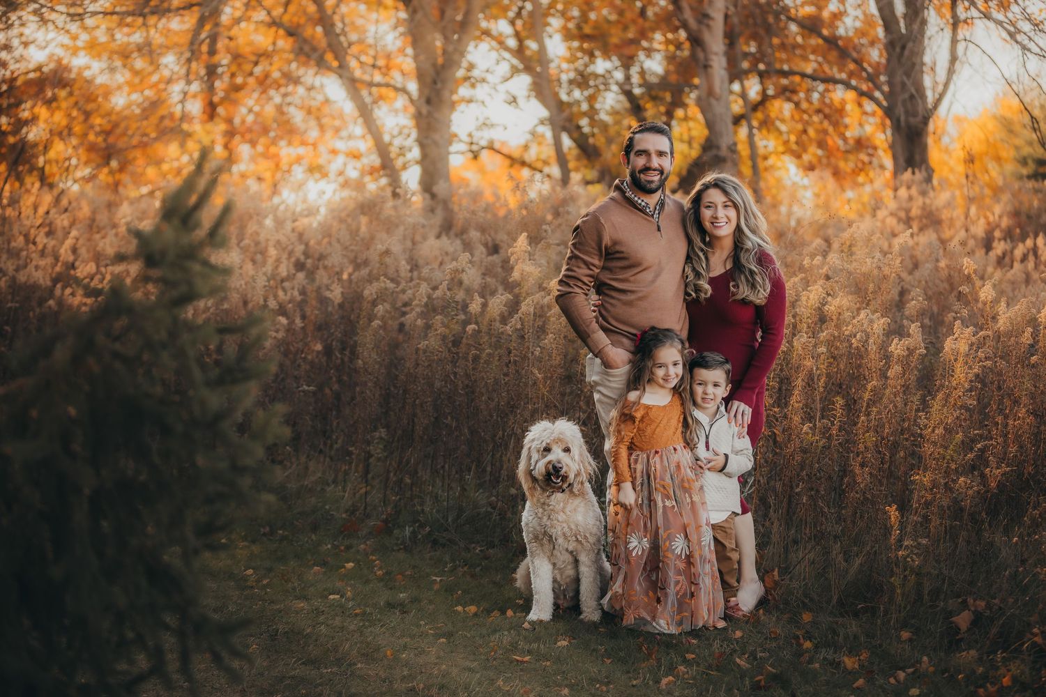 A group poses for autumn family photos in a wooded area with golden fall foliage and warm sunset lighting.