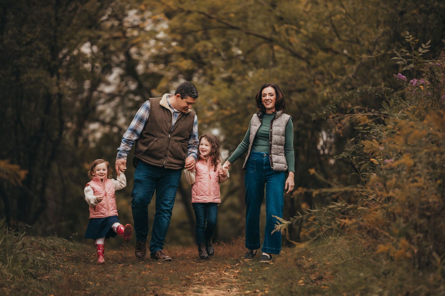 Family walks together through autumn forest path with fallen leaves and golden light filtering through trees.
