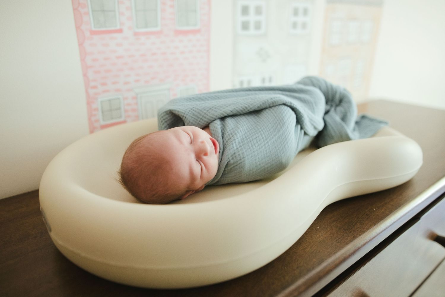 Newborn baby swaddled in blue wrap sleeping peacefully in a white ergonomic lounger on a wooden dresser.