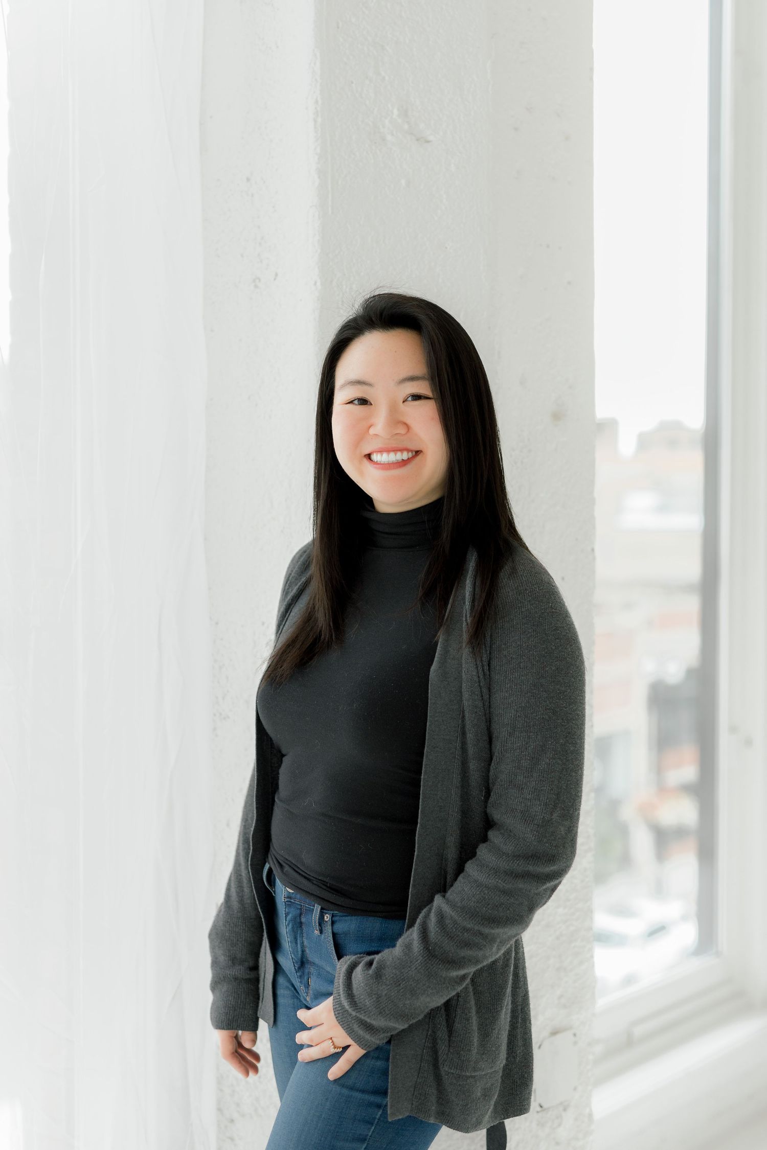 Person in black turtleneck and gray cardigan smiles in front of white walls.