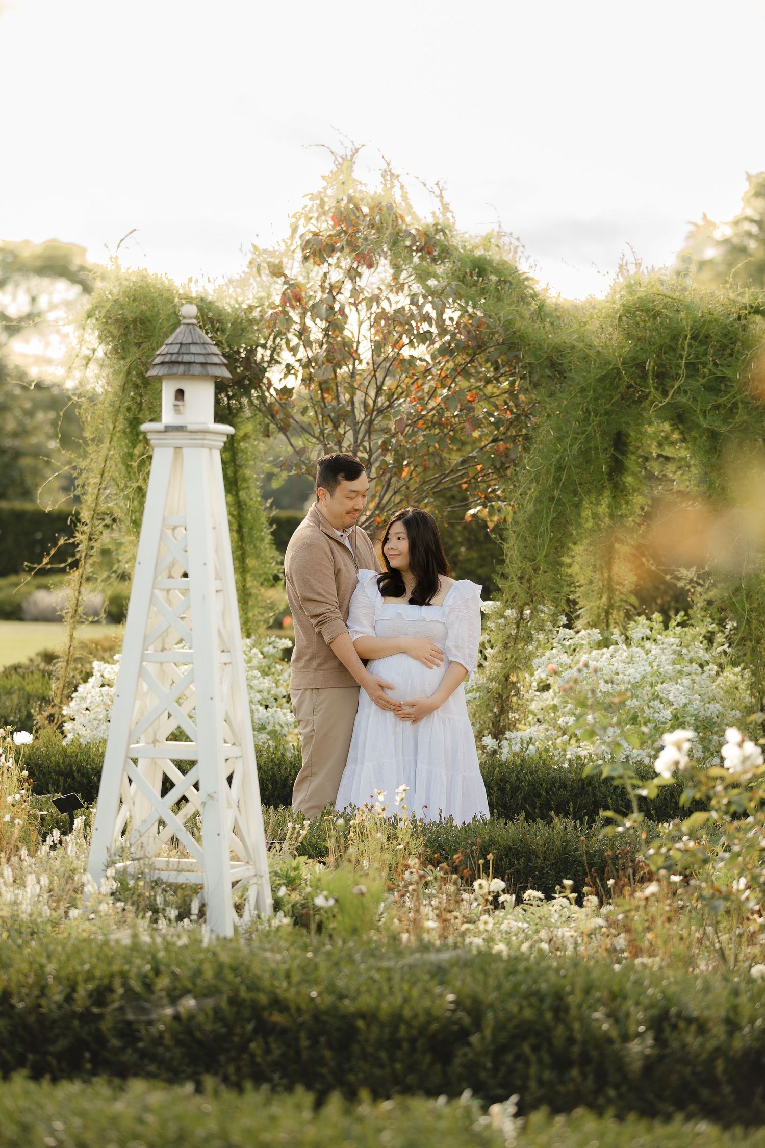 A romantic maternity photoshoot in a garden with white lighthouse decorations and blooming flowers.