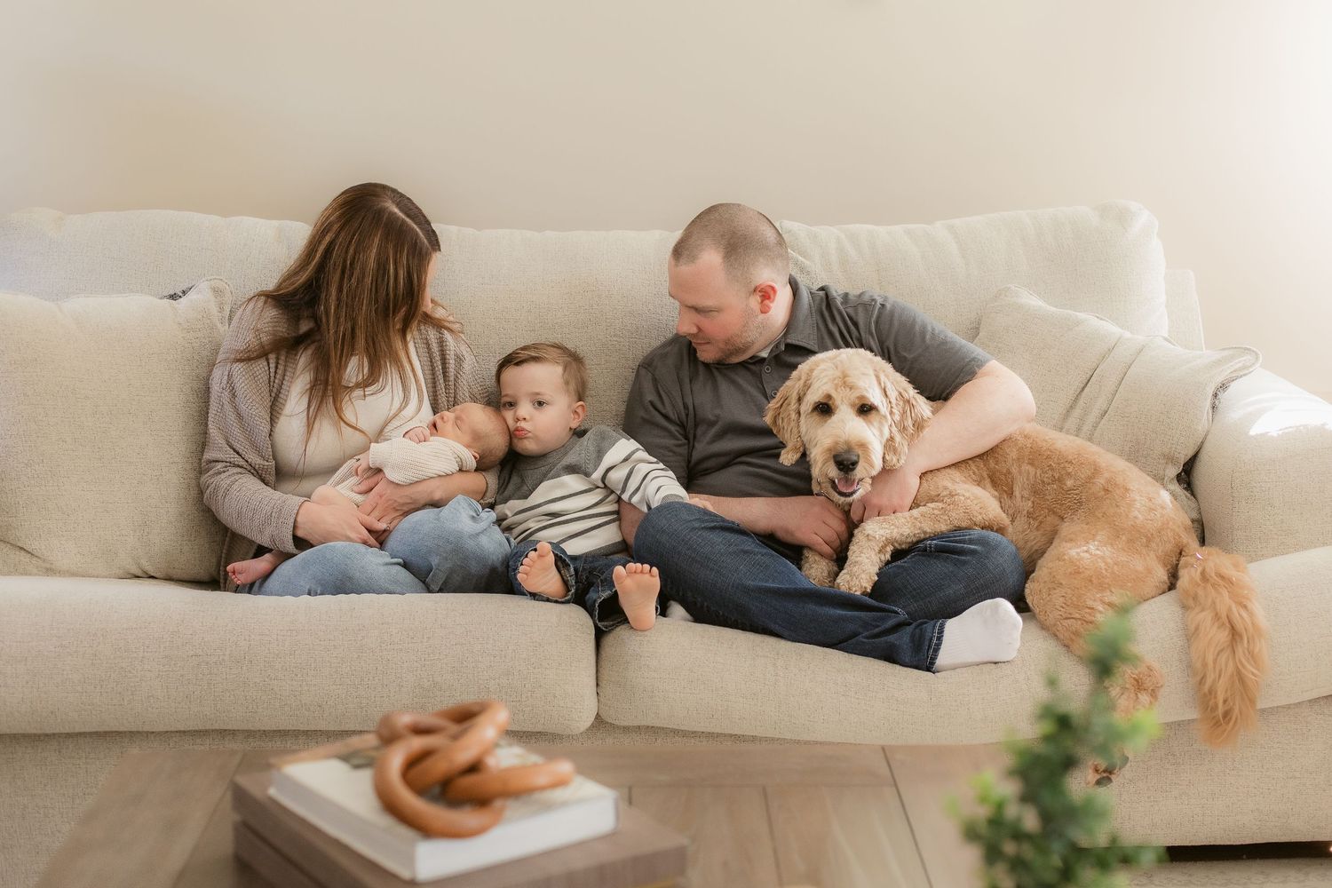 Family relaxing on beige couch with golden retriever and snacks on nearby table.