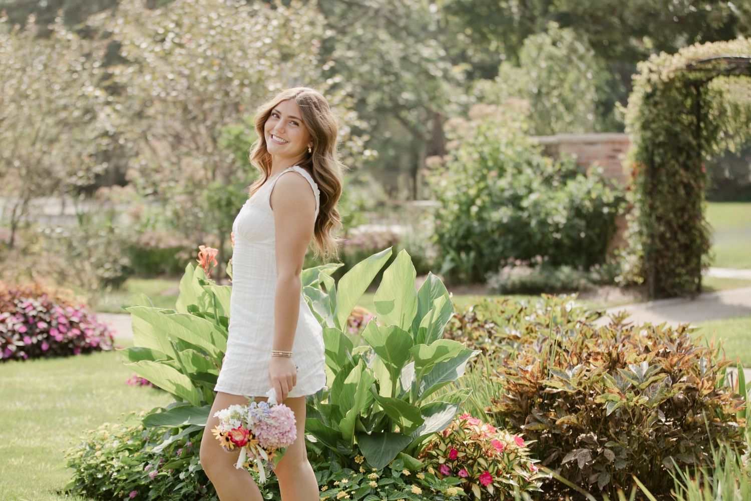 Woman in white dress standing among lush tropical garden plants, smiling over shoulder on sunny day.
